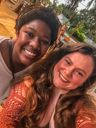 Two smiling friends taking a selfie on a wooden outdoor patio deck with warm sunset lighting, trees and backyard seating visible in the background.