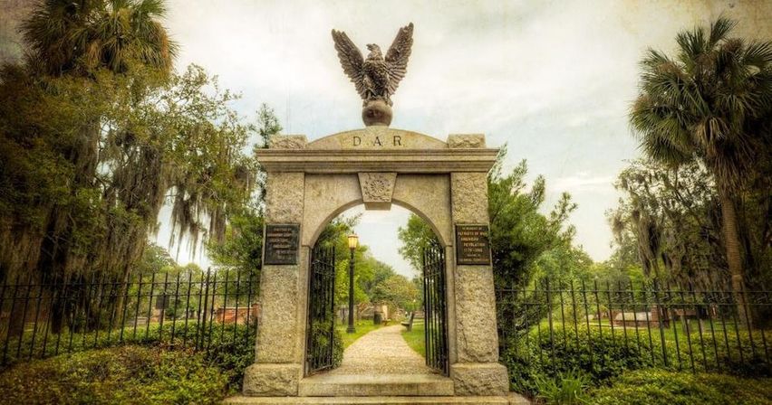 Granite archway topped by an eagle statue with open iron gates leading to a winding path through moss-draped oaks and palms at a historic cemetery entrance.