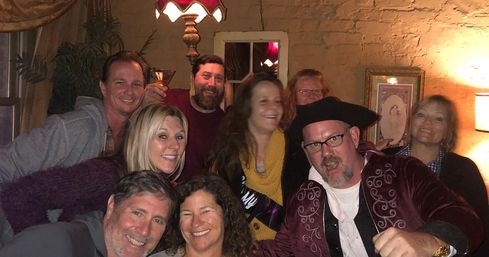 Smiling group of friends in a cozy dimly lit lounge with exposed brick and vintage lamps, celebrating — man in a pirate-style hat and a visible cocktail.