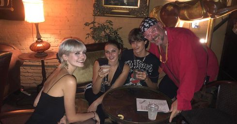 Four friends smiling in a cozy, dim brick-walled cocktail lounge gathered around a small round wooden table with drinks, a vintage lamp and decorative elephant statue in the background.