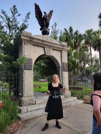 Stone DAR arch topped by an eagle statue marking a tree-lined historic cemetery entrance with palms and Spanish moss; person in a black dress and boots gesturing in the foreground.