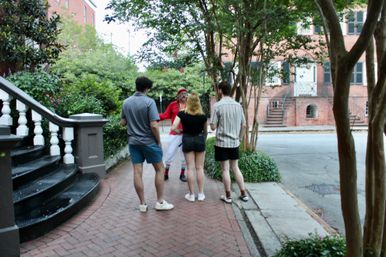 Four people chatting on a tree-lined brick sidewalk by historic red-brick row houses and stoops, casual summer clothes and a man in a red bandana gesturing during a daytime stroll.