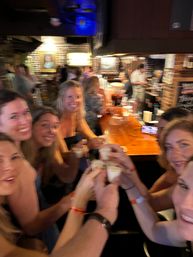 Group of friends clinking shot glasses and smiling at a lively brick-walled neighborhood bar over a wooden counter during a night out.