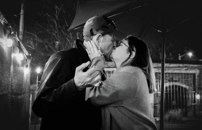 Black-and-white photo of a couple sharing a kiss beneath string lights on an outdoor patio at night — woman in glasses and sweater, man wearing a camo cap.
