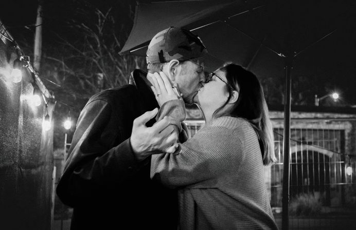 Black-and-white photo of a couple sharing a kiss beneath string lights on an outdoor patio at night — woman in glasses and sweater, man wearing a camo cap.