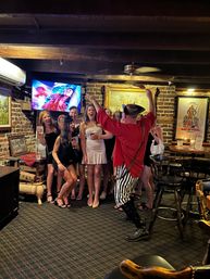 Group of women laughing and posing in a cozy brick-walled pub as a lively costumed performer in a red shirt and striped pants leads the celebration under wooden beams, framed art, and a TV screen.