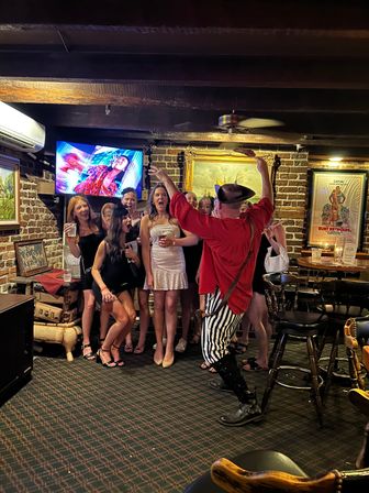 Group of women laughing and posing in a cozy brick-walled pub as a lively costumed performer in a red shirt and striped pants leads the celebration under wooden beams, framed art, and a TV screen.