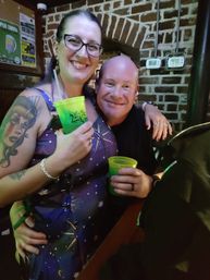 Smiling friends at a cozy brick-walled pub holding neon green cups, woman with glasses and a sleeve tattoo enjoying nightlife.