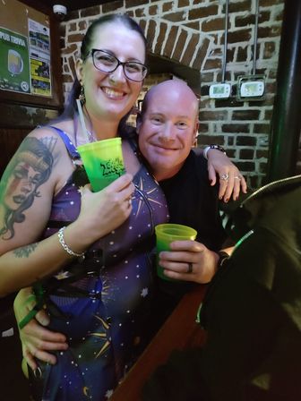 Smiling friends at a cozy brick-walled pub holding neon green cups, woman with glasses and a sleeve tattoo enjoying nightlife.