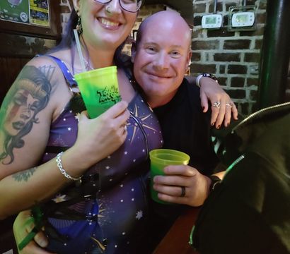 Smiling friends at a cozy brick-walled pub holding neon green cups, woman with glasses and a sleeve tattoo enjoying nightlife.