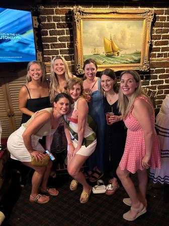 Seven women in summer dresses smiling and posing for a night-out photo inside a cozy brick-walled bar, holding drinks in front of a large ornate framed sailboat painting.