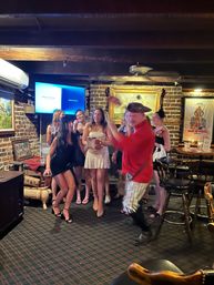 Lively, cozy brick-walled bar with exposed wooden beams; a group of women in dresses laugh and cheer while a man in a red shirt and pirate-style hat dances near bar stools, framed art, and a TV screen.