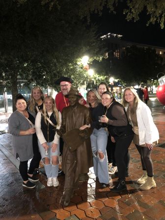 Smiling group of friends posing around a bronze statue of a hat‑wearing man in a wet downtown plaza at night, holiday lights and a giant red ornament in the background.