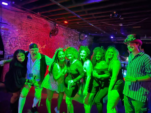 Eight friends smiling and posing in neon green and purple lights at an underground bar/nightclub, wearing party outfits and costumes and holding drinks in front of an exposed brick wall.