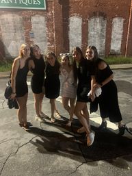 Six friends in dresses posing for a nighttime group photo on a downtown street in front of a historic brick storefront with an antiques sign; one wearing a white sash and novelty sunglasses for a bachelorette celebration.