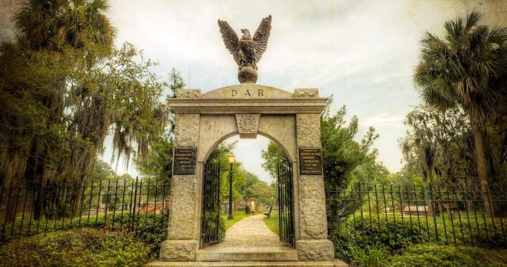 Stone archway cemetery entrance topped by an eagle statue, iron gates opening to a tree-lined path with oak trees draped in Spanish moss.