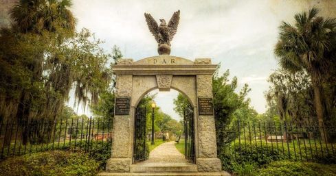 Stone archway cemetery entrance topped by an eagle statue, iron gates opening to a tree-lined path with oak trees draped in Spanish moss.