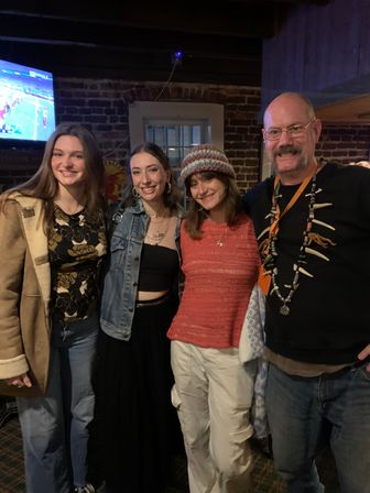 Four smiling adults posing together inside a cozy brick-walled pub — three women in casual layered outfits and a man wearing a beaded necklace, with a TV showing sports in the background.