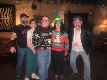 Group of five adults at a holiday party in a cozy brick‑walled lounge, wearing festive ugly sweaters and an elf hat, posing and smiling under warm ambient lighting.