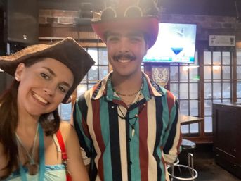 Cheerful selfie of two people in costume hats — woman in a tricorn hat and man in a red cowboy hat and striped shirt inside a cozy pub with brick wall, wooden windows and a TV screen in the background.