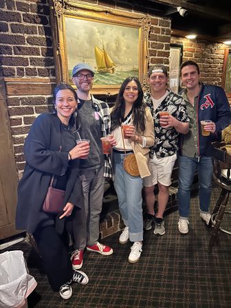 Five friends smiling with drinks in a cozy brick-walled pub, standing beneath a framed sailboat painting — casual night out