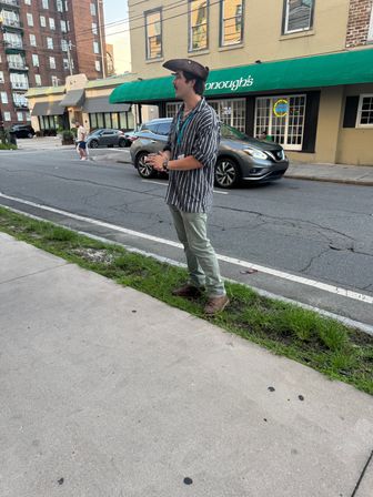 Person wearing a tricorn hat and striped shirt gestures while standing on the curb outside a green-awning storefront on a city street with parked cars.