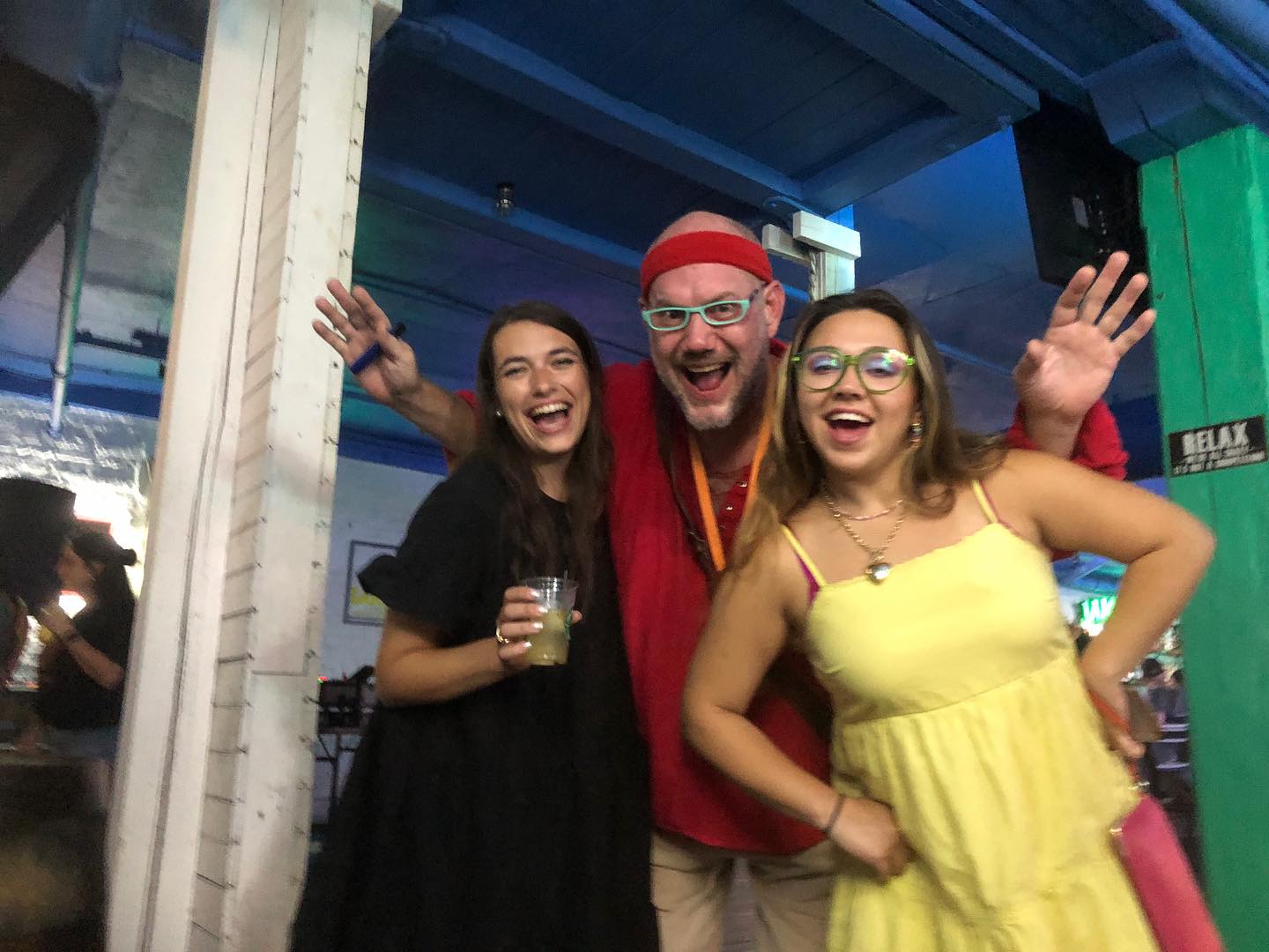 Three happy people posing at a lively indoor bar/event space — a man in a red headband and teal glasses between two women, one holding a cocktail and the other in a yellow sundress.