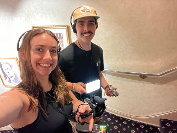 Two people wearing headphones smile for a selfie in a carpeted stairwell, holding a camera with shotgun microphone, LED light panel and a portable audio recorder, framed art on the wall.