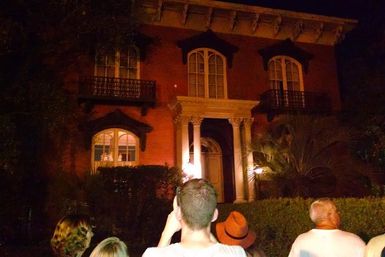 Tour group gazing at a lit historic red-brick mansion at night with columned entrance, arched windows, second-floor balconies and palm landscaping