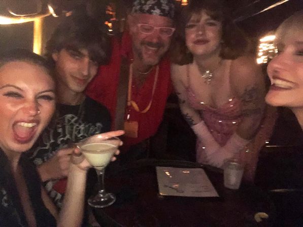 Five friends laughing at a dimly lit bar, toasting with a cocktail glass in a lively nightlife scene with warm string lights.