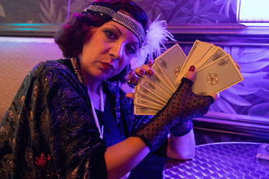 Fortune-teller in 1920s flapper costume with feathered headband, pearls and lace gloves holding a fanned deck of tarot cards at a purple-lit parlor table