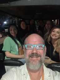 Nighttime group selfie aboard an open-sided golf cart on a city street — smiling friends with a bearded man wearing bright blue glasses in front.