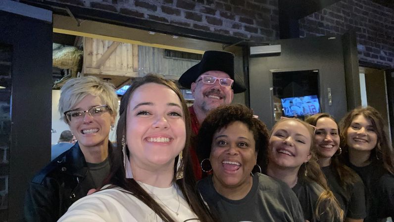 Seven-person group selfie of smiling adults on a casual night out in a cozy brick-walled bar or restaurant, people clustered close together with one wearing a black cowboy hat and glasses, lively social atmosphere.