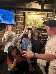 Five people striking surprised poses in a cozy brick-walled pub beneath a vintage sailboat painting, tattoos and a cocktail visible.