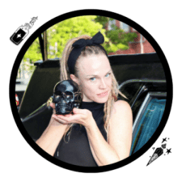 Person in a black headband and sleeveless top holding a glossy black skull prop, posed outdoors by a parked car in an urban setting