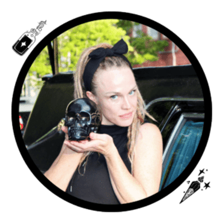 Person in a black headband and sleeveless top holding a glossy black skull prop, posed outdoors by a parked car in an urban setting