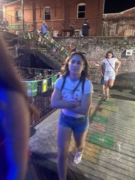 Young woman in a white t-shirt and denim shorts walking toward the camera through a lively outdoor bar alley at night, with brick walls, a metal staircase and railings hung with beer banners and people in the background.