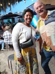 Smiling woman in a white top and yellow floral skirt with a black crossbody bag on a lively outdoor patio at dusk under string lights, a blurred man holding a drink leans in while a seated crowd and performer appear in the background.