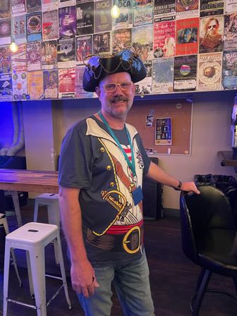 Smiling man wearing glasses, a decorative pirate hat and a pirate-print t-shirt, standing by bar stools in a lively indoor music venue with colorful concert posters on the wall and hanging lights.