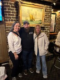 Three smiling adults in casual clothes posing together inside a cozy brick-walled pub beneath a framed sailboat painting.