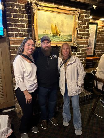 Three smiling adults in casual clothes posing together inside a cozy brick-walled pub beneath a framed sailboat painting.