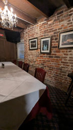 Cozy private dining room with a long white-clothed table, wooden chairs, exposed brick wall with framed cityscape photos, and a crystal chandelier overhead.
