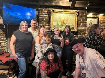 Group of friends smiling and posing in a cozy brick-walled, nautical‑themed pub with a framed sailboat painting, TV screen, and drinks.