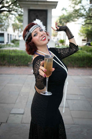 Playful woman in 1920s flapper costume — black lace dress, long pearls, feathered headband and lace gloves — offering a champagne toast in an outdoor city plaza.