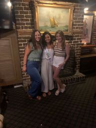 Three women posing for a group photo in a dimly lit brick-walled pub beneath a framed sailboat painting, wearing casual summer outfits and a celebratory sash for a night out.
