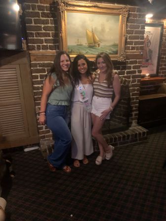 Three women posing for a group photo in a dimly lit brick-walled pub beneath a framed sailboat painting, wearing casual summer outfits and a celebratory sash for a night out.