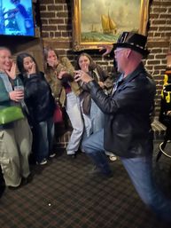 Performer in a top hat and leather jacket playfully miming a magic trick to a laughing group of friends inside a cozy brick-walled pub with a framed sailboat painting.