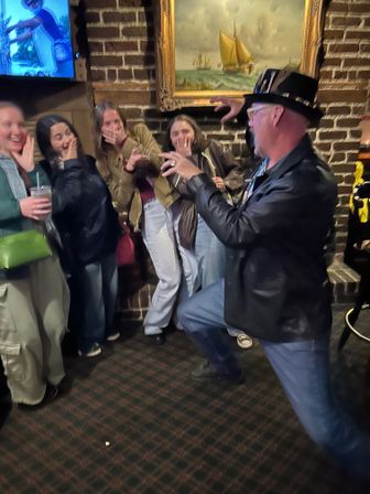 Performer in a top hat and leather jacket playfully miming a magic trick to a laughing group of friends inside a cozy brick-walled pub with a framed sailboat painting.