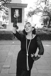 Black-and-white photo of a woman in a 1920s flapper outfit — lace dress, long pearl rope, feathered headband and fingerless gloves — raising a champagne flute in a celebratory toast in an outdoor urban plaza.