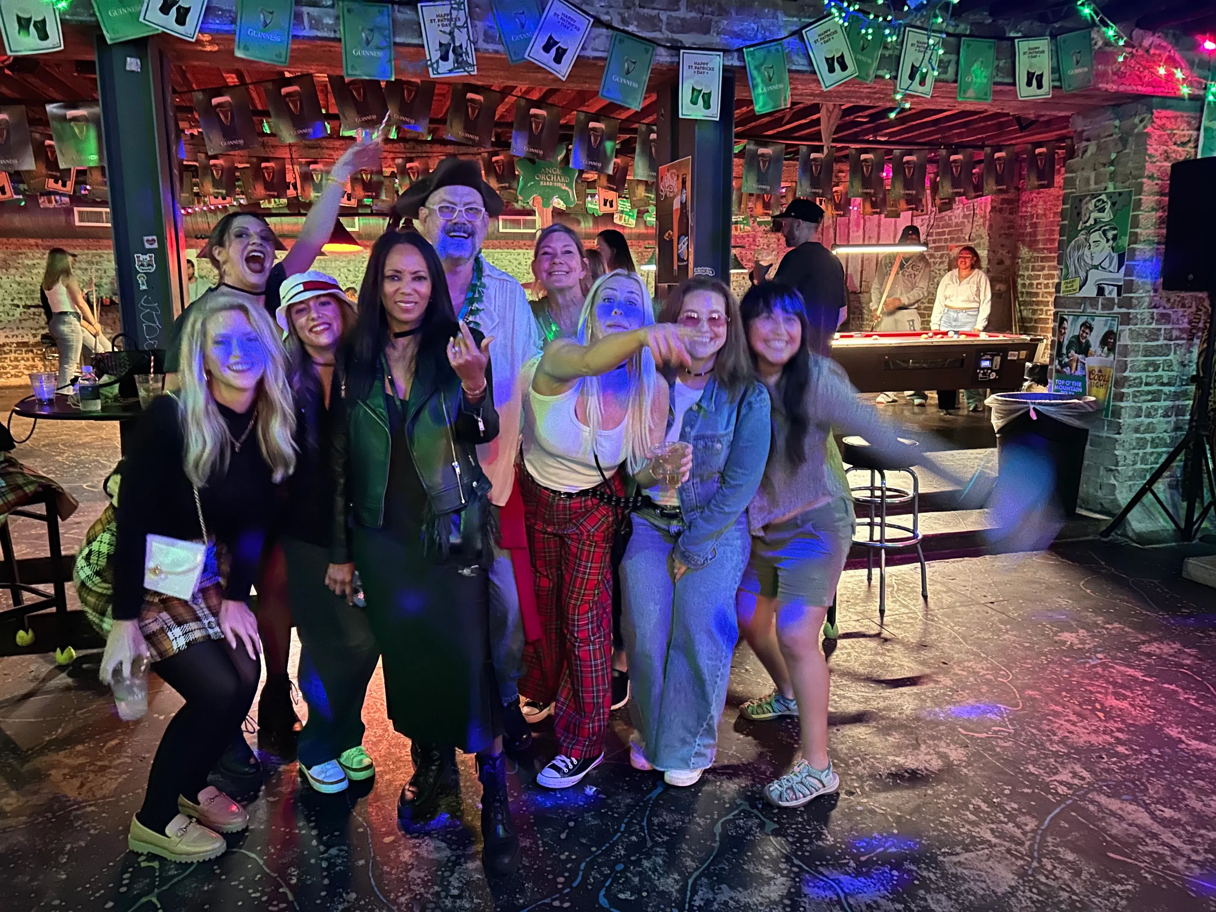 Group of friends enjoying a night out in a lively urban bar with exposed brick walls, hanging green banners and string lights, colorful disco lighting, and a pool table in the background.